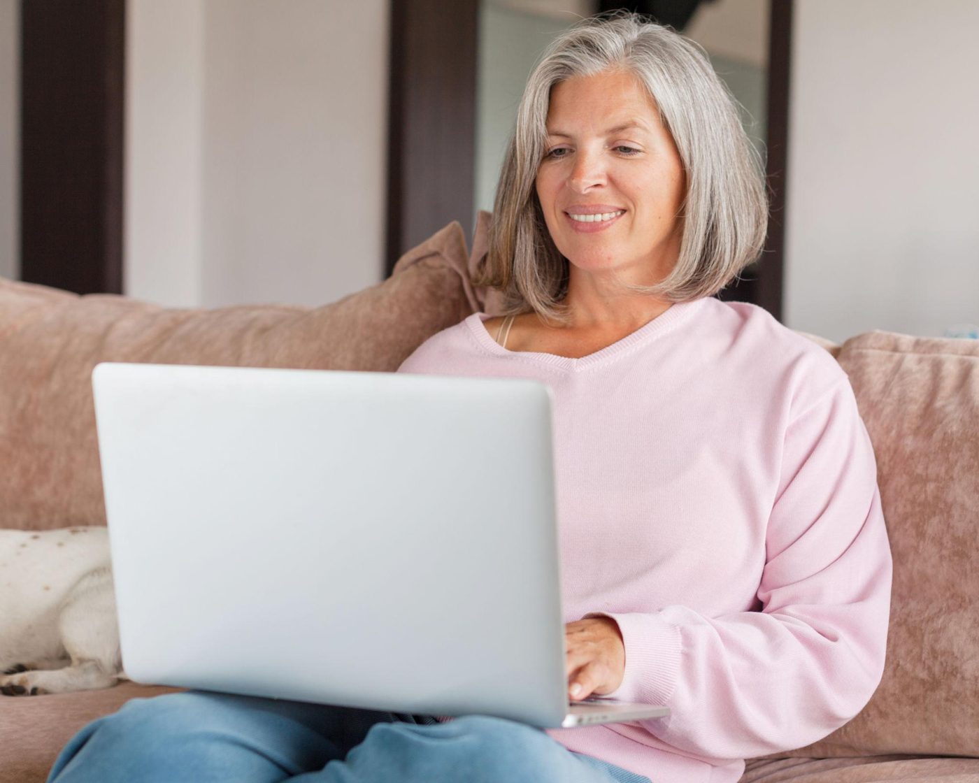 A smiling woman with grey hair relaxing on her sofa with her laptop